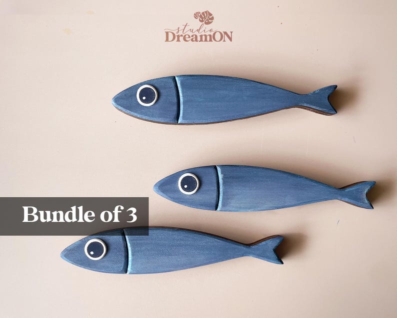 May include: Three wooden fish decorations in varying shades of blue. Each fish has a white and black eye and a detailed tail fin. The text "Bundle of 3" is displayed in a black box. The Studio DreamON logo is in the top right corner.