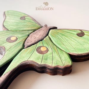 K&ouml;nnte beinhalten: Eine Wanddekoration aus Holz, die einen gr&uuml;nen Luna-Motte mit braunen Akzenten zeigt. Der Falter hat ein detailliertes Design mit Adern auf seinen Fl&uuml;geln und gelben Flecken.