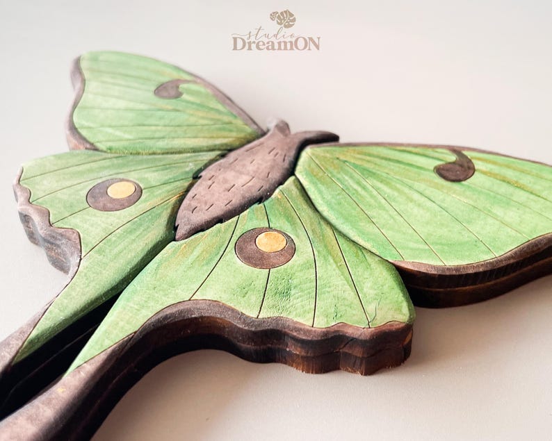 K&ouml;nnte beinhalten: Eine Wanddekoration aus Holz, die einen gr&uuml;nen Luna-Motte mit braunen Akzenten zeigt. Der Falter hat ein detailliertes Design mit Adern auf seinen Fl&uuml;geln und gelben Flecken.