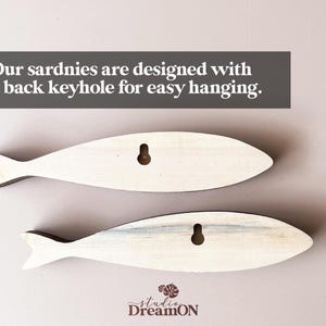 May include: Two wooden fish-shaped wall decorations. The fish are light-colored with a keyhole on the back for hanging. The text on the image says, "Our sardnies are designed with a back keyhole for easy hanging."