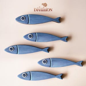 May include: Five blue wooden fish decorations with white and black eye details. The fish are arranged in a diagonal pattern against a beige background. The fish have a simple, stylized design, with a fin at the tail end. The top of the image has the text "studio DreamON".