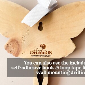 K&ouml;nnte beinhalten: Eine h&ouml;lzerne Schmetterlings-Wanddekoration mit nat&uuml;rlicher Holzoptik. Der Schmetterling hat oben ein Loch zum Aufh&auml;ngen und der Text "studi DreamON www.dreamon.STUDIO" ist unten eingraviert. Sie k&ouml;nnen auch das mitgelieferte selbstklebende Haken- und Schleifenband f&uuml;r die Wandmontage verwenden.