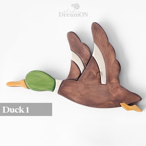 K&ouml;nnte beinhalten: H&ouml;lzerne Enten-Wanddekoration in Braun, Gr&uuml;n und Wei&szlig; bemalt. Die Ente ist im Flug mit ausgebreiteten Fl&uuml;geln. Der Text "Duck 1" befindet sich unter der Ente.