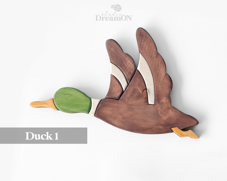 K&ouml;nnte beinhalten: H&ouml;lzerne Enten-Wanddekoration in Braun, Gr&uuml;n und Wei&szlig; bemalt. Die Ente ist im Flug mit ausgebreiteten Fl&uuml;geln. Der Text "Duck 1" befindet sich unter der Ente.