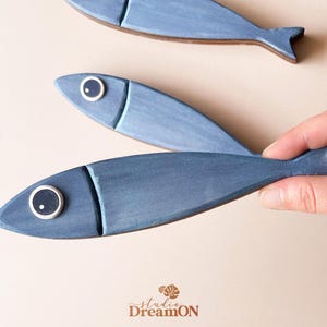 May include: Three blue wooden fish decorations. One fish is being held by a hand. The fish have a painted design with a lighter blue head and a white and black eye. The fish are a decorative item.