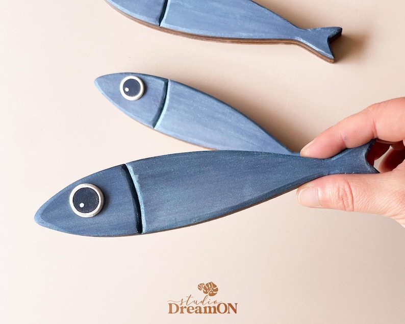 May include: Three blue wooden fish decorations. One fish is being held by a hand. The fish have a painted design with a lighter blue head and a white and black eye. The fish are a decorative item.