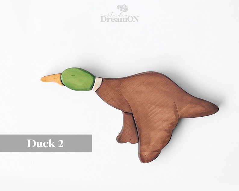K&ouml;nnte beinhalten: Ein h&ouml;lzernes Entenspielzeug, das in Braun-, Gr&uuml;n- und Gelbt&ouml;nen bemalt ist. Die Ente ist im Flug mit ausgebreiteten Fl&uuml;geln. Das Spielzeug ist mit "Ente 2" beschriftet.