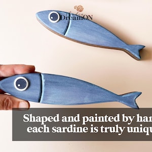 May include: Two blue wooden sardine-shaped decorations with white and black eye details. One is held by a hand. The text on the image reads: "Shaped and painted by hand, each sardine is truly unique."