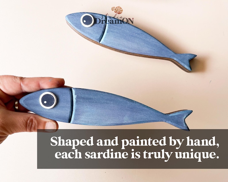 May include: Two blue wooden sardine-shaped decorations with white and black eye details. One is held by a hand. The text on the image reads: "Shaped and painted by hand, each sardine is truly unique."