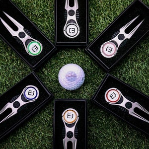 Pode incluir: Seis ferramentas de reparo de divots de bola de golfe prateadas em caixas pretas com círculos de cores diferentes e as letras "EJ" no centro. Uma bola de golfe branca está no centro da imagem.