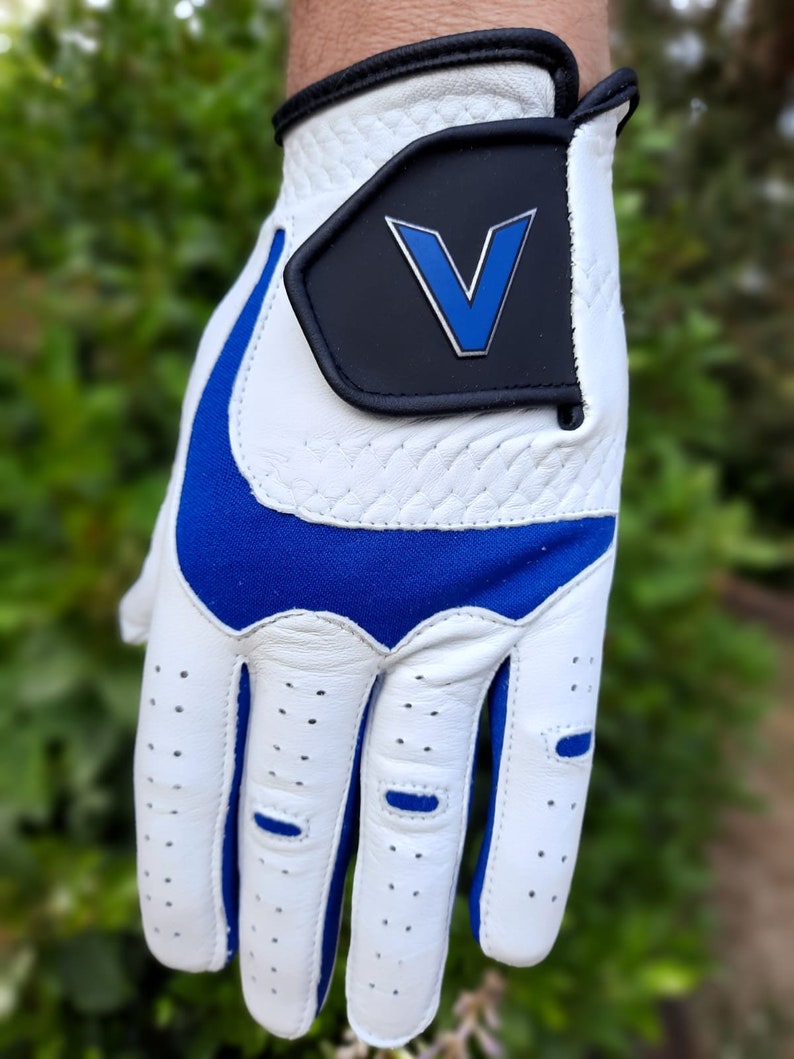 Op de afbeelding: Witte golfhandschoen met een blauwe accent en een zwarte patch met een blauw "V" logo.