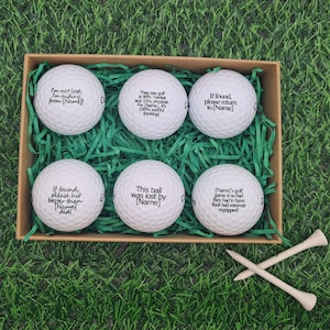 Op de afbeelding: Een set van zes witte golfballen, elk bedrukt met een humoristische tekst in het zwart, gepresenteerd in een bruine doos met groen papier. Twee houten golf tees zijn gekruist aan de onderkant. De golfballen hebben zinnen als "Indien gevonden, graag terug naar [Name]"