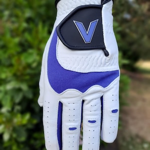Op de afbeelding: Witte golfhandschoen met een paarse palm en een zwarte patch met een blauwe "V" op de achterkant van de hand.