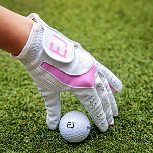 Może przedstawiać: Biała rękawica golfowa z różowymi akcentami i inicjałami "EJ" na pasku nadgarstka. Biała piłka golfowa z inicjałami "EJ" znajduje się na pierwszym planie.