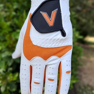 Op de afbeelding: Witte golfhandschoen met oranje accenten en een zwarte patch met een witte "V" op de achterkant van de hand.
