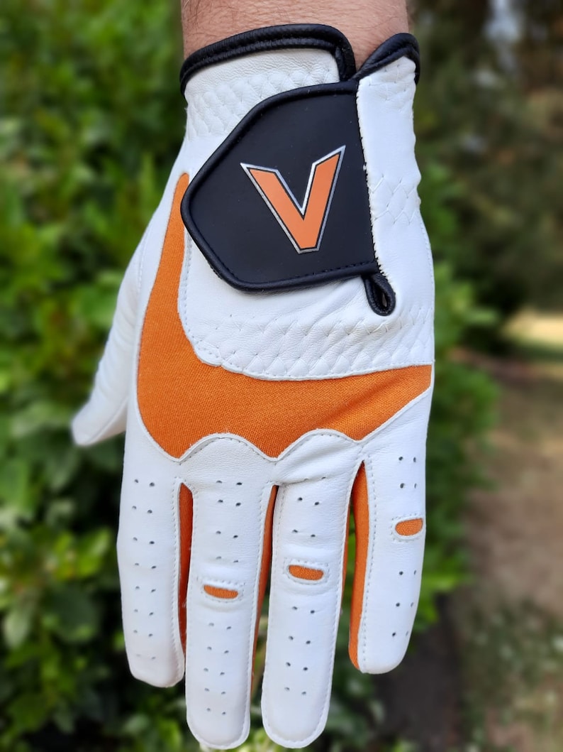 Op de afbeelding: Witte golfhandschoen met oranje accenten en een zwarte patch met een witte "V" op de achterkant van de hand.