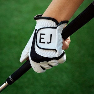 May include: White golf glove with black accents and the initials "EJ" on the strap. The glove is worn on a hand holding a golf club.