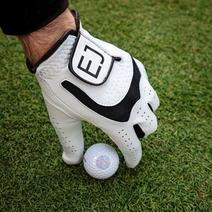 Puede incluir: Un guante de golf blanco con un diseño de patrón blanco y negro. El guante tiene un cierre de velcro con la letra "E" en él. Una bola de golf blanca está siendo sostenida por el guante.