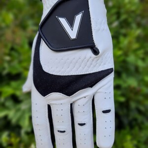 Op de afbeelding: Witte golfhandschoen met een zwart V-logo op de achterkant van de hand. De handschoen heeft een zwart-wit ontwerp op de palm en vingers.