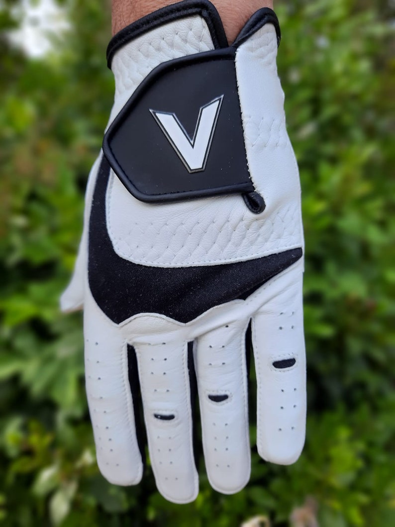 Op de afbeelding: Witte golfhandschoen met een zwart V-logo op de achterkant van de hand. De handschoen heeft een zwart-wit ontwerp op de palm en vingers.