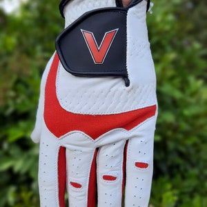 Op de afbeelding: Witte golfhandschoen met rode accenten en een zwarte patch met een rode "V" op de rug van de hand.