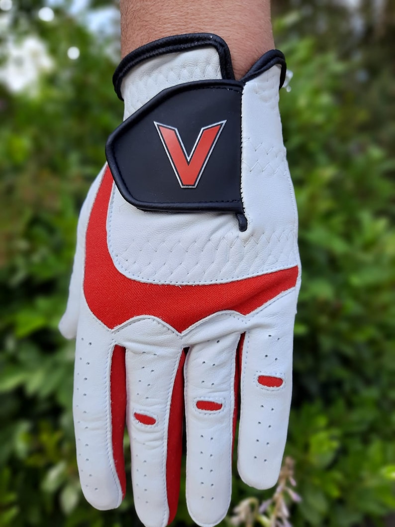 Op de afbeelding: Witte golfhandschoen met rode accenten en een zwarte patch met een rode "V" op de rug van de hand.