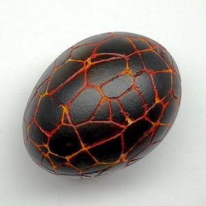 May include: A decorative egg with a cracked, textured surface. The egg is primarily black with a network of red and orange lines, resembling lava or a fiery pattern. The egg shape is oval.