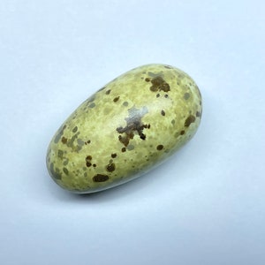 以下が含まれることがあります： 薄い緑色のベースカラーと茶色の斑点がある、楕円形の卵。卵の表面は滑らかな質感です。卵は自然の産物で、おそらく鳥のもので、白い背景に置かれています。