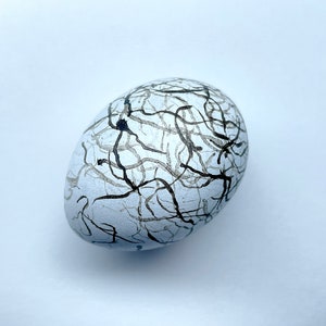 May include: A white, egg-shaped decorative stone with a network of black and gray lines painted across its surface. The lines create a web-like pattern, giving the stone a unique, artistic appearance. The stone is set against a plain white background.