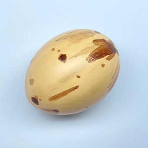 May include: A decorative egg with a light tan base colour and brown brushstroke patterns. The egg-shaped object has a smooth surface and is painted with irregular brown spots and lines, creating a unique design.