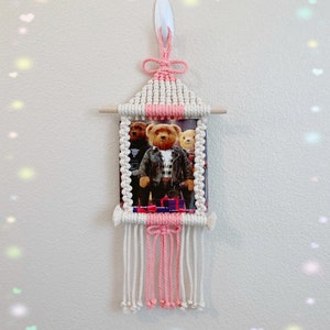 May include: A pink and white macrame wall hanging with a picture of three teddy bears wearing clothes. The wall hanging has a wooden dowel at the top and a pink and white tassel at the bottom.