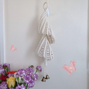 May include: A white macrame wall hanging with three tiers of woven shells and a white ring at the top. The hanging has two small gold bells at the bottom.