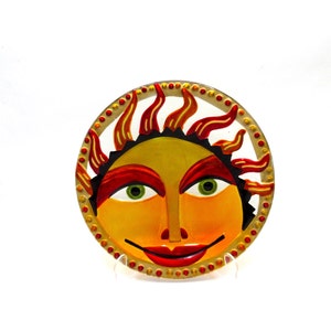 May include: A round, hand-painted sun face with a smiling expression. The sun has a yellow face, red and orange flames, and green eyes. The border is gold with red dots.