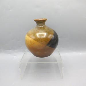 May include: A small, round ceramic vase with a narrow neck and a flared rim. The surface features a speckled, multi-toned glaze with sections of golden yellow, warm brown, and a deep, glossy black color block on the side, creating a smooth finish.