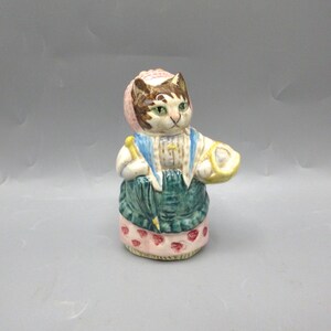 May include: A ceramic figurine of a cat dressed in a bonnet, blue vest, and green skirt. The cat holds a yellow basket and a closed umbrella. The base of the figurine is pink with red heart-shaped designs.