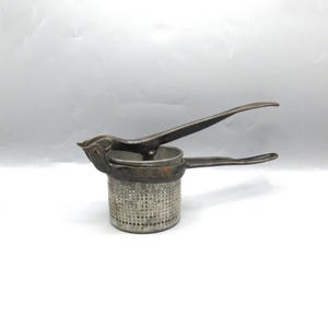 May include: Vintage potato ricer made of metal. The ricer has a perforated bowl and long handles for pressing potatoes. The metal has a worn, aged appearance, suggesting it is an antique kitchen tool. The design is simple and functional.