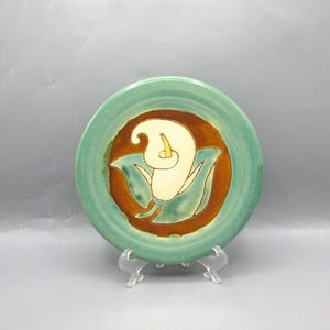 May include: A decorative ceramic plate with a hand-painted calla lily design. The plate features a turquoise rim, a brown center, and a white lily with green leaves. The plate is displayed on a clear plastic stand.