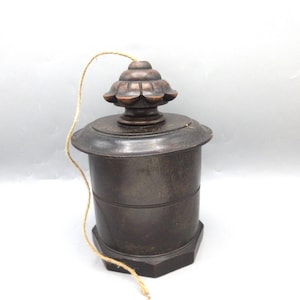 May include: A dark brown, wooden container with a decorative lid and a hexagonal base. The lid has a floral design and a string is attached. The container has a cylindrical body with horizontal grooves. The object is set against a white background.