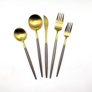 May include: A set of gold and gray flatware, including a knife, fork, and two spoons. The handles are gray and the metal parts are gold.