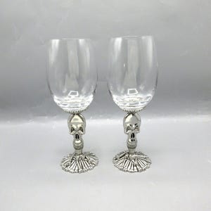 May include: A pair of clear glass wine glasses featuring silver-toned metal stems shaped like human skulls. The base of each glass is designed to resemble a collection of scattered human bones, providing a stable foundation for the stem.