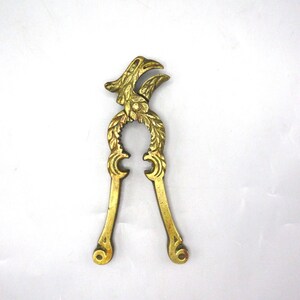 May include: A vintage brass nutcracker with an eagle head design. The nutcracker features a detailed eagle head at the top, a circular opening, and long handles. The brass has a warm, golden hue, and the design is ornate.