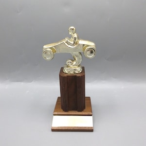 May include: A gold-colored trophy featuring a racer driving a go-kart. The trophy is mounted on a wooden base with a blank plaque.