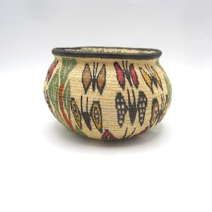 May include: A round, woven basket made of light tan natural fibers, featuring a repeating pattern of butterflies in red, yellow, brown, and black. The basket has a thick, dark brown rim and a rounded, bulbous shape with a wide opening at the top.