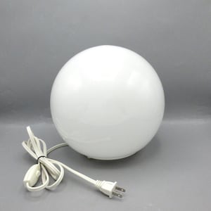 May include: A spherical white glass table lamp with a smooth, opaque finish. The lamp features a white electrical cord with a small inline toggle switch and a standard two-prong plug attached to the base of the globe-shaped light fixture.