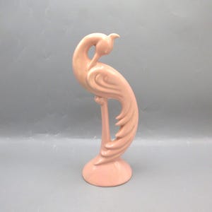 May include: A ceramic figurine of a stylized bird in a soft pink hue. The bird has a long, curved neck and a detailed tail, perched on a circular base. The figurine is a decorative item, likely for display on a shelf or table.