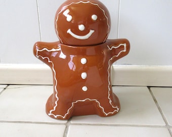 Puede incluir: Un tarro de cerámica marrón para galletas con forma de hombre de jengibre. El tarro presenta detalles de glaseado blanco pintados en la cabeza, los brazos y las piernas, junto con tres botones circulares blancos en el centro del torso y una cara sonriente.