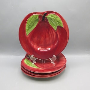 May include: A set of red ceramic plates shaped like apples, with green leaf accents. The plates are stacked, with one plate upright, showcasing the apple design. The plates are on a light gray background.