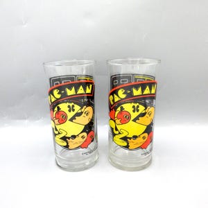 May include: Two clear glass drinking glasses with a Pac-Man design. The glasses feature Pac-Man and ghosts in yellow, orange, red, and blue.