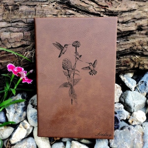 May include: A brown leather journal with a detailed engraving of hummingbirds and a flower. The journal is rectangular and has rounded corners. The name "Lindsey" is inscribed in the bottom right corner.