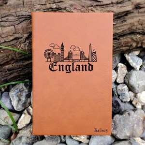 May include: A brown leather journal with a black outline of the London skyline and the word "England" engraved on the front. The name "Kelsey" is engraved at the bottom of the journal.